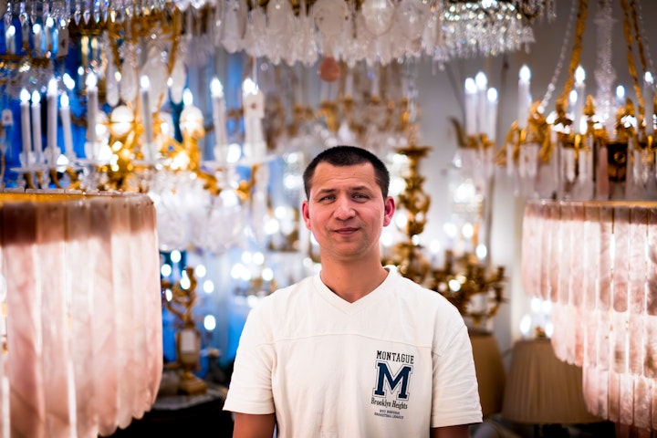 Open Britain: Portrait of a Diverse Nation - Dimitri Stefanov’s shop on the King’s Road, Chelsea is a ‘lighthouse’, only this one draws you in. When I photographed him in his shop, he was surrounded by friends and colleagues who he had just cooked for: His London family. It’s easy to see why, there is something extremely endearing about Dimitri.
Dimitri was born in Bulgaria. He didn’t go to school much. At fourteen he moved with his parents from Bulgaria to Greece and worked in polytunnels. Something he looks back at with great fondness.
In 2009 he moved to the UK to be with his friend an Antique Lighting dealer. The shift from aubergines to chandeliers was an easy one for Dimitri, despite absolutely no experience, he loved it. He helped with cleaning and maintenance of lights and running the shop.
Dimitri’s innate talent was spotted five years later by an octogenarian premier artisan chandelier maker called Philip Turner. Philip took him under his wing teaching him restoration, pinning and all the skills needed to create new and bring antique fittings back to life.
In 2010 Dimitri picked up some old iron chandelier frames that were being thrown out. He was ‘drawn’ to some raw rock crystals at the back of a friends chest of drawers and so the alchemy began. Three days later they were sold.
Soon after, encouraged by Philip, Dimitri left the shop to start making lamps of his own. The next chandeliers, a pair of swans, sold just as fast.
Dimitri is now one of the most pre-eminent rock crystal chandelier specialists in the world. He counts many of the world’s wealthiest people as his clients including celebrities, royalty, Claridge’s, Annabel’s and Gaudi Museum,Barcelona.
Dimitri is Philips legacy to the antique world and creating chandelier is Dimitri’s absolute passion. Having said that he still misses the polytunnels.