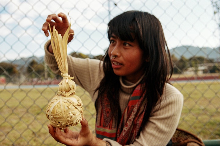 Amita is balancing a 'pash-pash' ball on top of 'maize ball-game' ball. Both are made from corn leaves. The 'pash-pash' ball has a tail and flies much like a shuttlecock. The tail mimics the growth of the plant, and is played in the autumn during the typical harvest of corn.