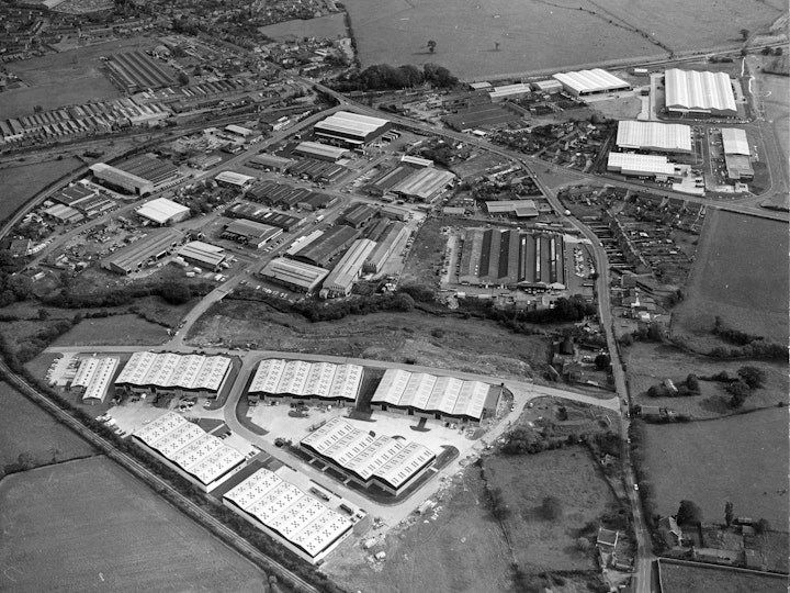 Scan of an original aerial photograph looking east of the various trading estates and railway network in Yate in 1974. 
The Beeches Industrial Estate is in the foreground to the north of the River Frome. Stover Trading Estate is in the centre flanked by Badminton Road in the east, Stover Road to the west (right in the photo) and North Road to the north (left on the photo).
Badminton Road Trading Estate is in the south (top right of image) which is sandwiched between Badminton Road (A432) and the main Gloucester to Bristol railway line that runs North - South (horizontally in the image). 
Westerleigh Common (A.K.A. Yate Common) before major tree planting and rewilding is to the south east (top right of the image). The rein or brook that drains the common can be seen diagonally crossing the common lined by bushes and the football pitches on the playing fields segment of the common can be seen top centre. 
Sunnyside Playing Field with football pitch, which was home to Yate Town Football Club at this time is seen adjacent to Westerleigh Common in the top centre of the image with Parnalls factory opposite to the north on the other side of Station Road (A432) with many of the original aircraft production facilities running adjacent to the railway line. 
The remnants of Yate Railway Station and the Goods Shed can be seen just the south east of the junction of Station Road and North Road. The Thornbury branch line can be seen looping round from Yate junction, over North Road and in the foreground of the image to the north of the very new looking Beeches Trading Estate. Newmans Factory can be seen in the extreme top right of the image north of the adjacent to Station Road with the carpark clearly visible. 
Finally, Westerleigh Road can just about be seen running horizontally across the entire top of the image.