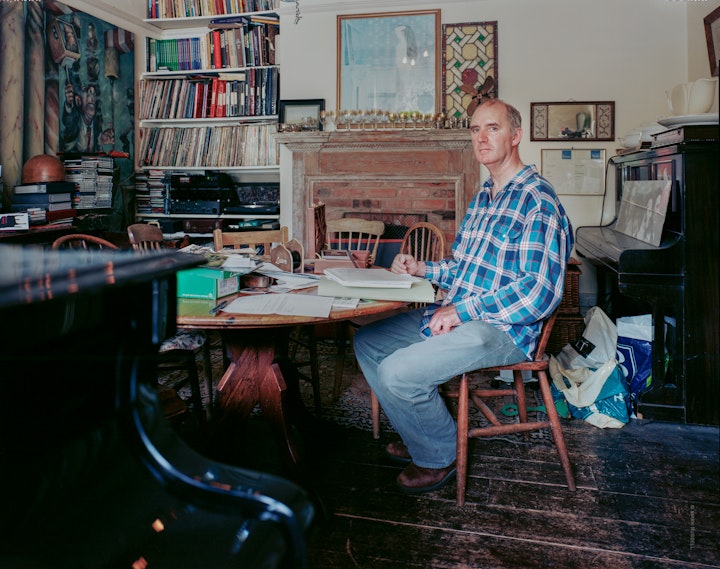 A portrait of Tim Andrews, curator of Over the Hill, a study of his journey with Parkinson's Disease. Tim sits at his table with paperwork. This was the the second photograph his photographic series.