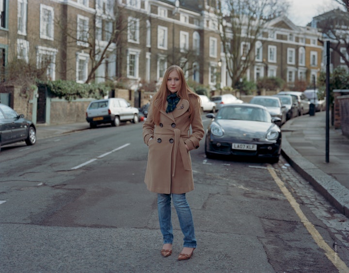 A portrait of Laura wearing a camel jacket and jeans stood in a crescent shaped road in Camden.