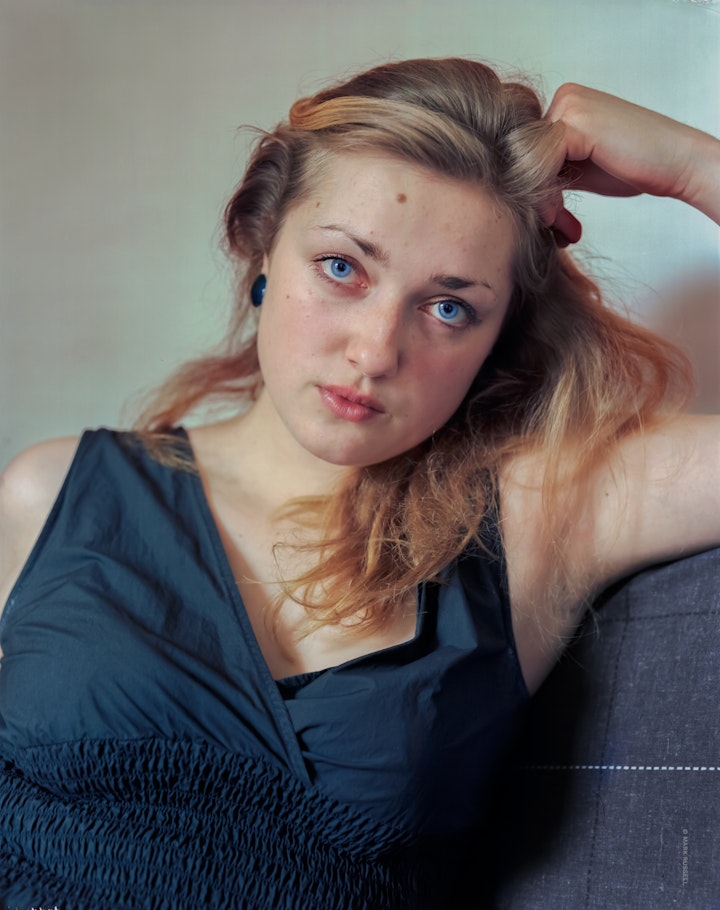 A portrait of Liis, a woman with clear blue eyes and blonde hair, sitting wearing a blue dress looking straight at camera.