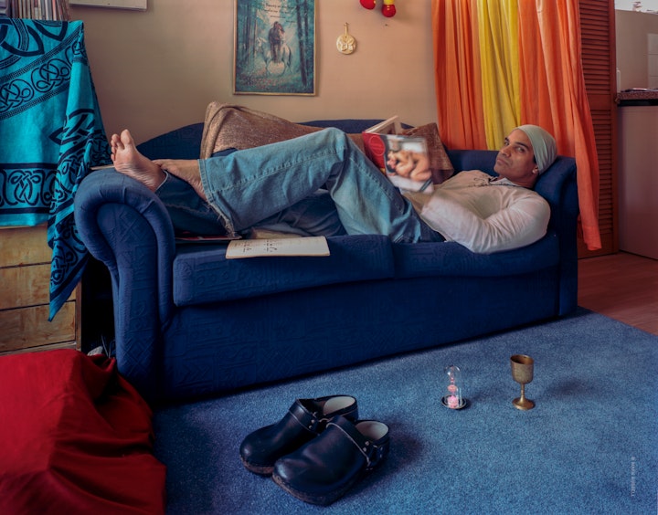 A portrait of Angel who is laying down on a small blue sofa in a colourful living room, reading the kama sutra with shoes and a small metal goblet in front on the floor.