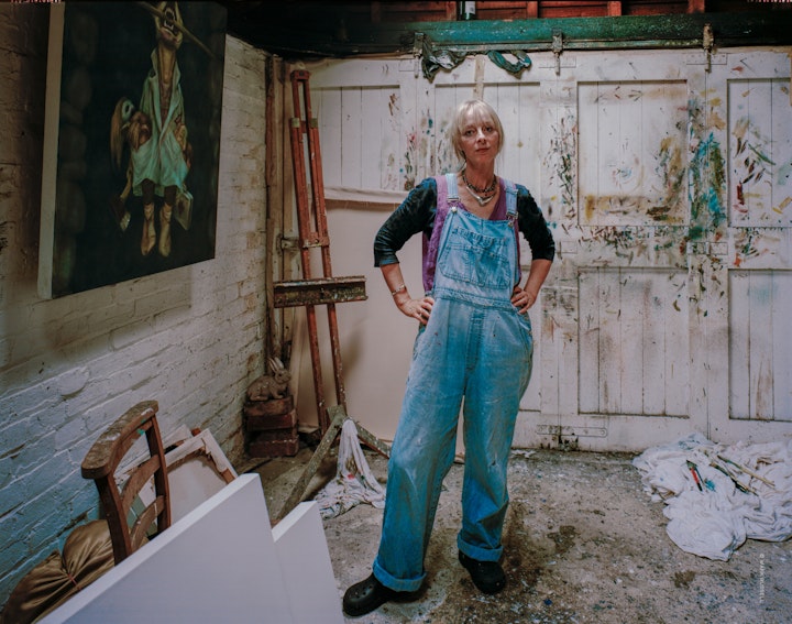 A portrait of the painter, Jane Andrews, standing in her studio surrounded by canvases, easels and walls marked with paintbrushes.