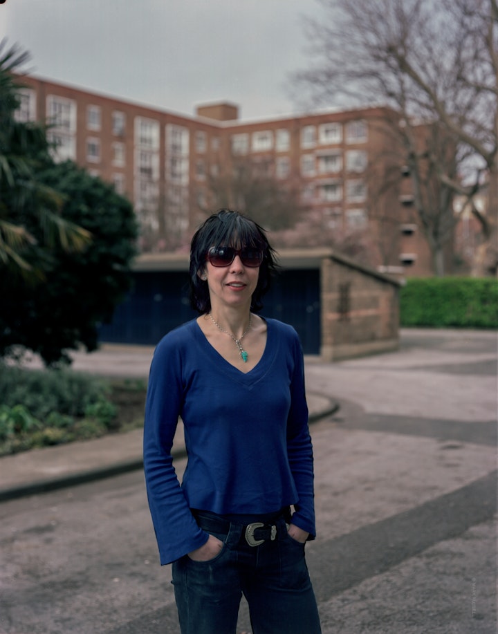 A portrait of Vicki, standing in the courtyard of a residential block in Clerkenwell, wearing dark sunglasses and a blue top.