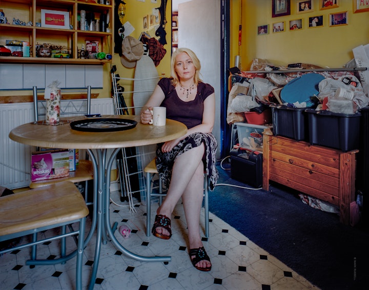A portrait of Tanya sitting cross-legged at her dining room table holding a mug of tea.