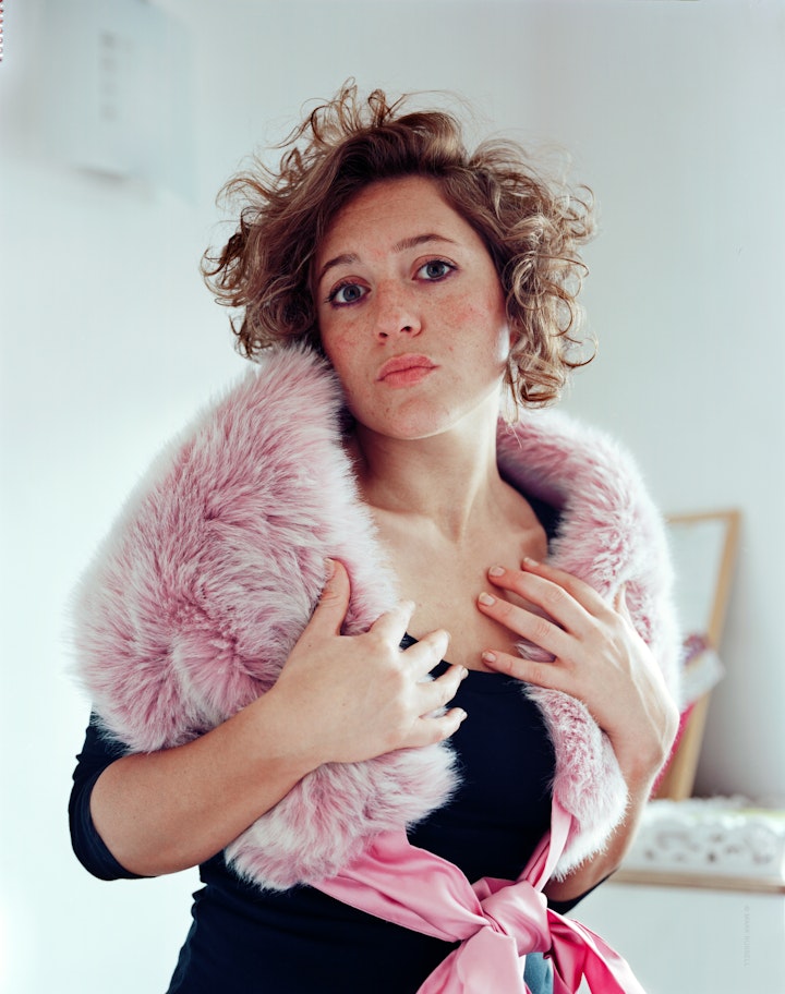 A portrait of Lucy wearing a pink father boa and a black dress, posing in her living room.