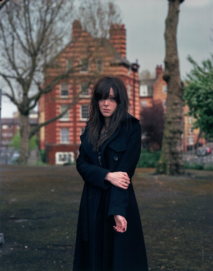 A portrait of Kirsty, wearing a long black coat with her arms across her body in front of a square in Shoreditch and a striped, red-brick building in the background.