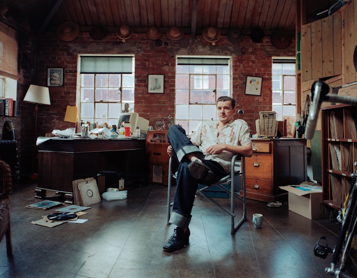 A portrait of Gordon, reclining in a chair in a large room which has a messy desk and a number of old vinyl records strewn over the floor.