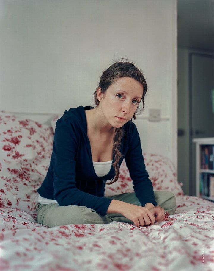 A portrait of Guida, a young woman sitting cross-legged on a double bed, leaning forward and looking into the camera.