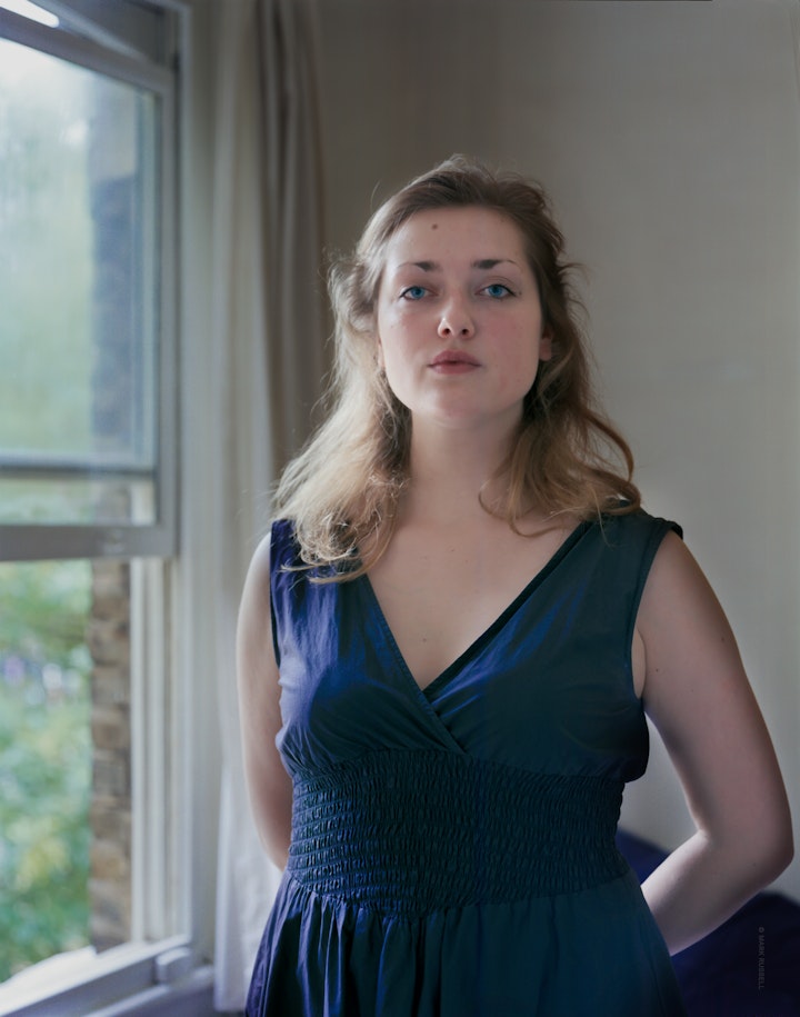 A portrait of Liis standing by the soft light of a window wearing a blue dress with her hands behind her.