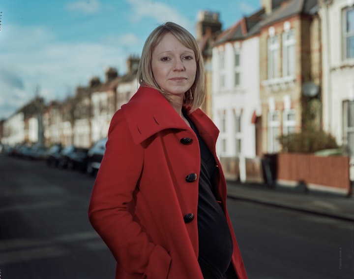 A portrait of Mel, three quarter length in a red coat looking straight at the camera with a London street behind her.
