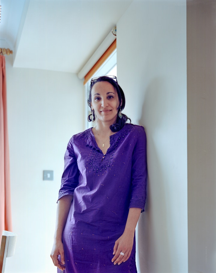 A portrait of Nadia, wearing a vibrant purple dress leaning against the wall in a brightly lit living room.