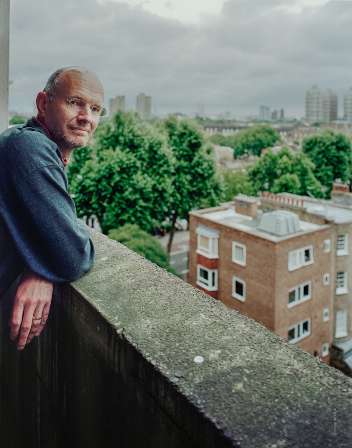 A portrait of author, Rob Pateman, standing at the top floor balcony of a tower block in south London looking at the camera with the city in the background.