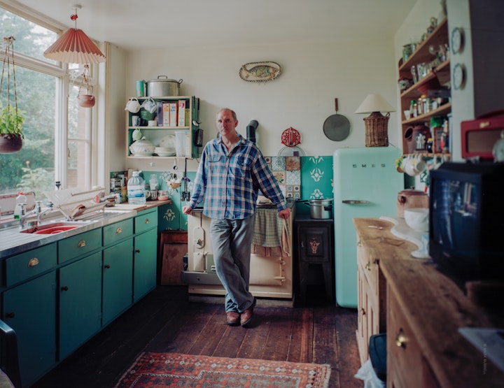 A portrait of Tim Andrews, curator of Over the Hill, a study of his journey with Parkinson's Disease. Tim stands by the range in his kitchen with soft sunlight from a garden window.
