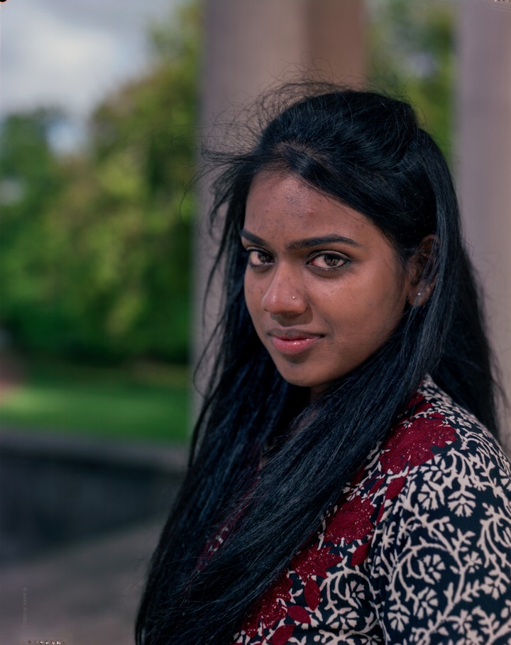 A portrait of Debrashree, standing looking at the camera with long black hair with the garden of Syon Park in the background.
