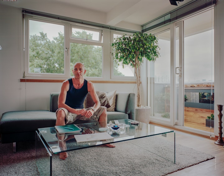 A portrait of Simon, sitting beyond a glass table in his living room with sunlight entering the room from an open balcony door.