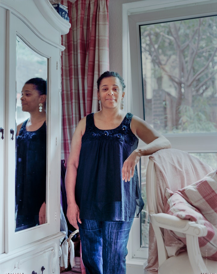 A portrait of Chris, standing in her bedroom, looking straight at camera with her reflection in a mirrored wardrobe.