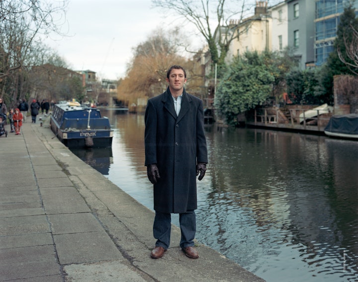 A portrait of Chris, standing by a canal in Camden wearing a business coat and leather gloves.