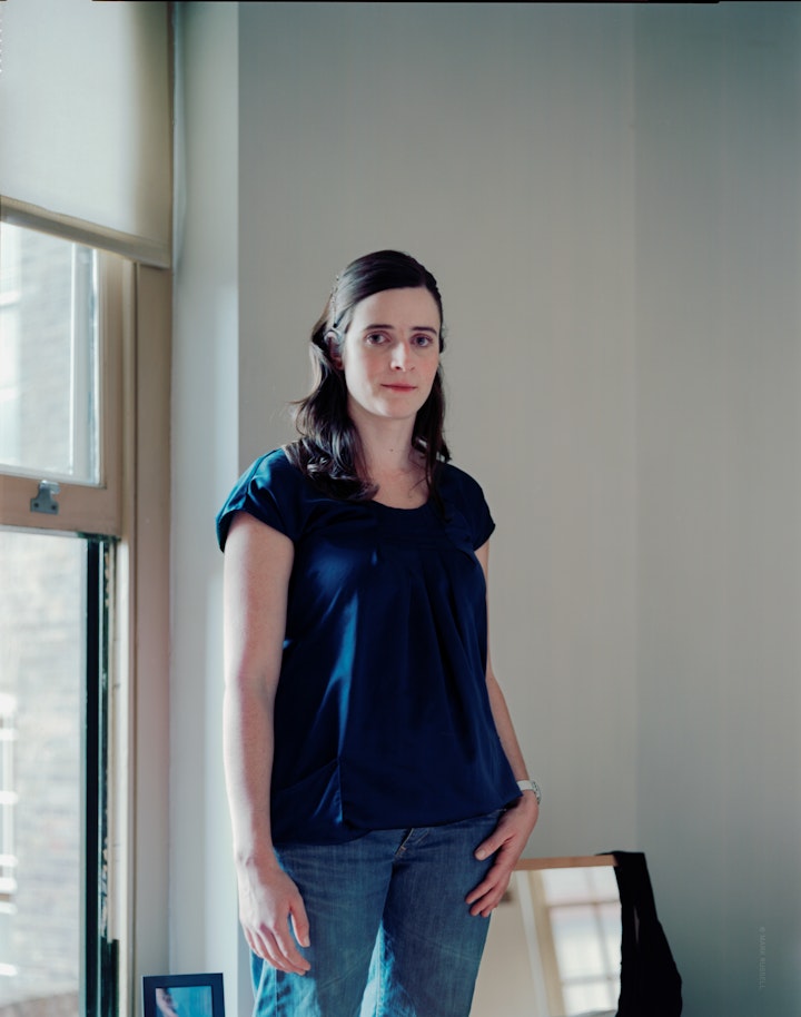 A portrait of Ana, wearing a blue shirt and jeans, standing beside a large window and a mirror.