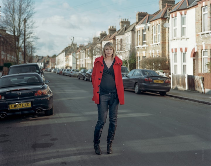 A portrait of Mel, who is pregnant, standing in an empty residential street with parked cars.