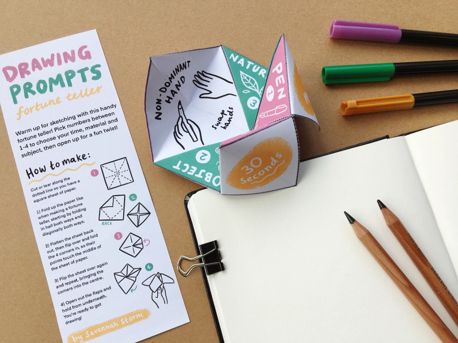 A table with an open sketchbook, three marker pens and two pencil lying next to it, and a paper fortune teller open with a drawing of hands. Text says 'Non-dominant hand drawing'