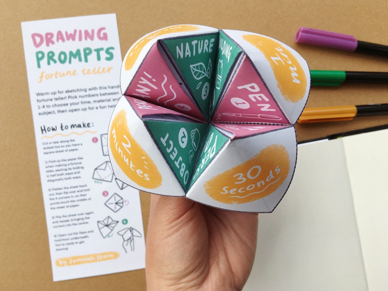 A hand holds up a printed paper fortune teller, with yellow circles on the outside, and green and pink sections inside.