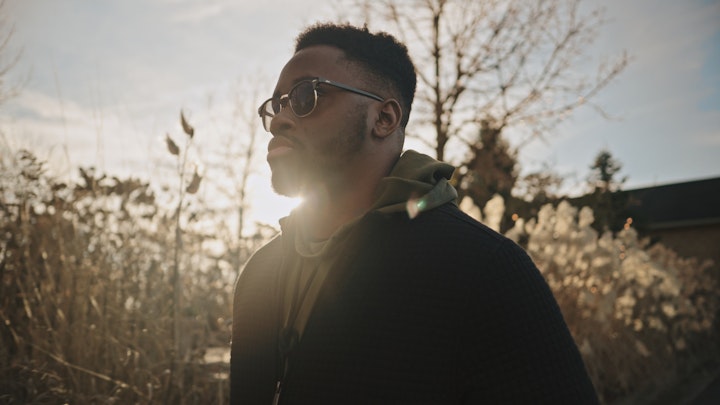 a man wearing sunglasses walking past bushes with golden sunlight streaming behind him