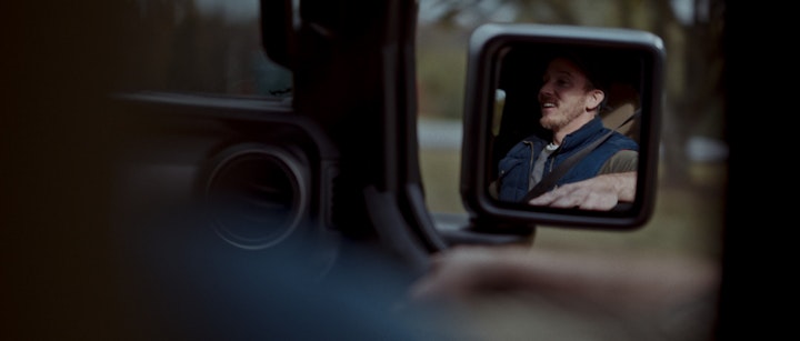 A man's reflection in a jeep's side mirror, riding and driving down the road