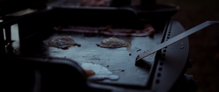 A grill with bacon and eggs sizzling, cooking, on it, with spatula scraping and flipping food
