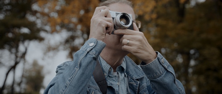 a man in a jean jacket taking a photo with an old camera, fall foliage in background