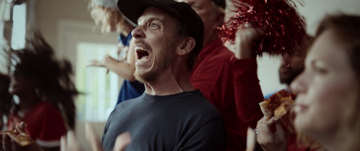 A man holding a pizza, cheering for a sports team and standing next to a cheerleader, both smiling and looking happy.