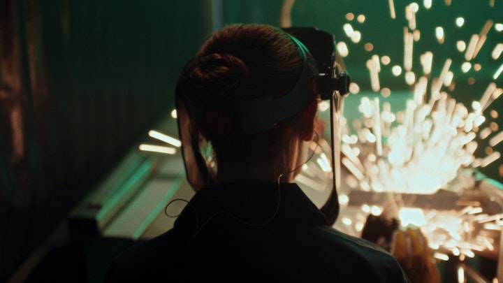 A woman in a helmet working on a machine. Sparks fly in a dark industrial shop on metal.