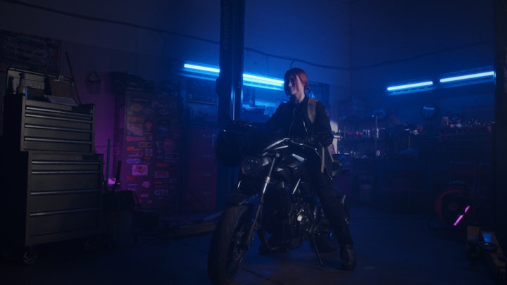 A woman sitting on a motorcycle in a pink and blue neon garage, ready to drive into the night and city.