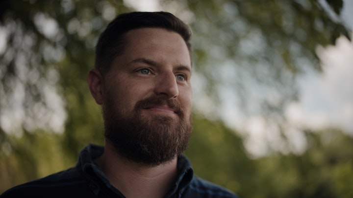 a man with a beard smiling and looking into distance green trees sway behind him
