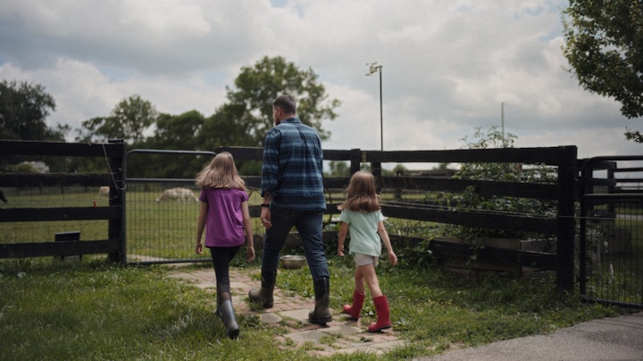 father walking with daughters on a farm toward fence with horses in background
