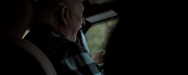 An older man driving a car, focused on the road ahead, with a determined expression on his face.