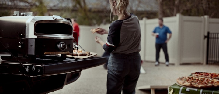 A woman standing near a pizza oven at a tailgate party and friends play cornhole in background yard