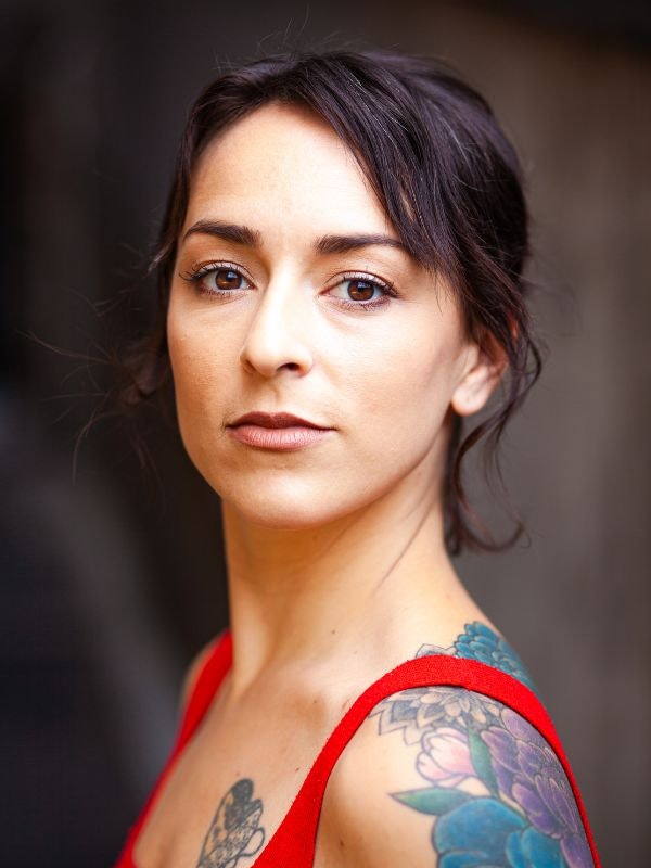 Danika Ciullo has a red tank top on with a colourful floral tattoo on her left shoulder. She faces side to the camera and looks into the lens, with brown hair tied up in the back.