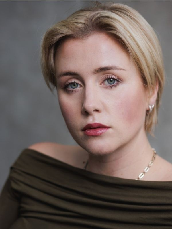 Connor Amor-Bendall wears a brown off the shoulder top, with red lipstick and piercing blue eyes. Blonde highlighted hair is cut just above the ears and parted slightly to the side. Silver hoop earrings and silver chain necklace.