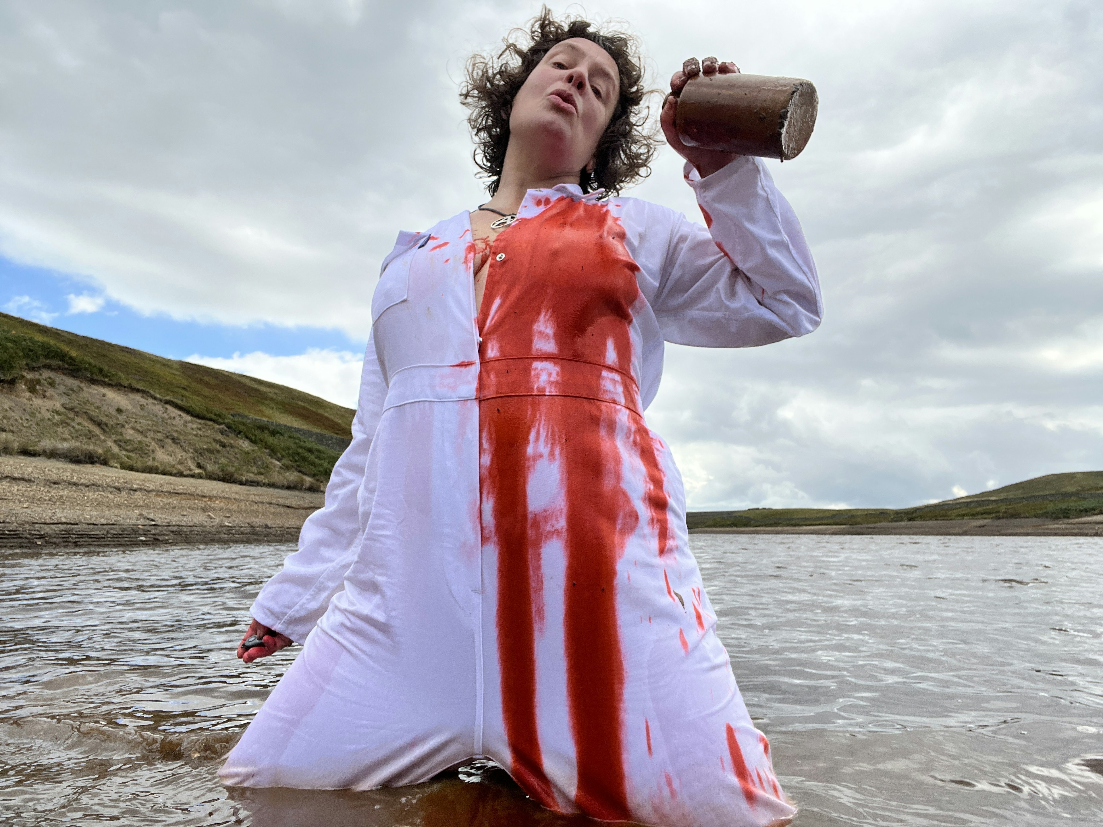 A woman kneels in the shallow of a lake, wearing white overalls she pours red ink over her heart. ‘A Type of Baptism’ by Alice Kin 2025